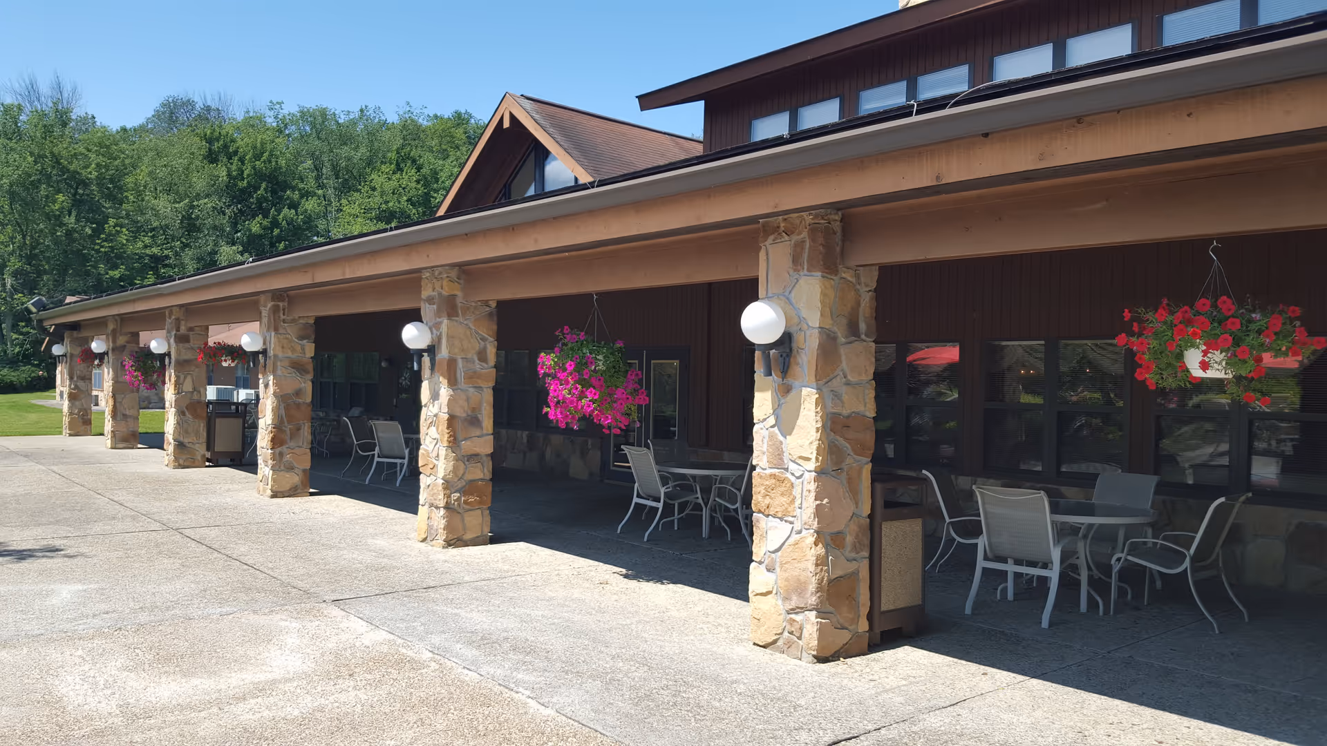 Outdoor covered patio area with stone pillars and hanging flower baskets. Several tables and chairs are arranged under the patio roof, with greenery and trees visible in the background under a clear blue sky.