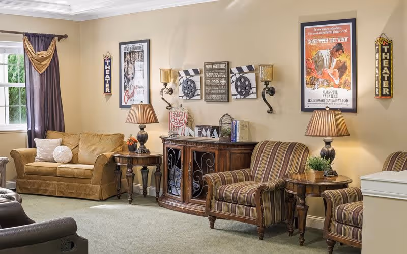 A cozy theater lounge featuring a brown sofa, striped armchairs, and decorative movie-themed wall art. The room has warm lighting and a welcoming atmosphere.