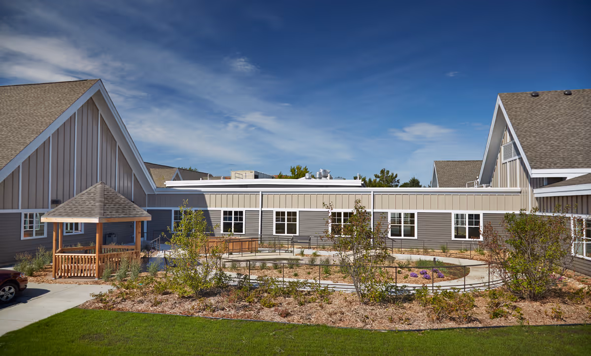 Exterior view of The Neighborhoods at Brookview facility showing a courtyard with a circular walking path, landscaped garden beds, small trees, and a wooden gazebo. The building has gray siding with white trim and multiple windows under a clear blue sky.