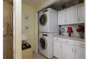 Stacked washer and dryer in a bright laundry area with white cabinets, countertop, and a view into a bathroom.