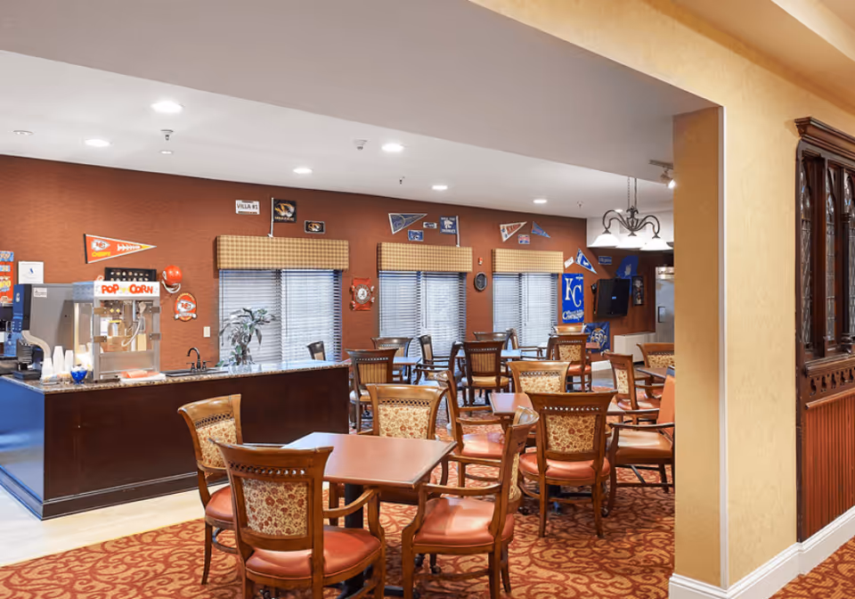 A dining area in a senior living facility with multiple wooden tables and chairs arranged on a patterned carpet. The walls are decorated with sports pennants and memorabilia. There is a counter with a popcorn machine and cups on the left side, and windows with blinds and valances along the back wall. The lighting is bright with recessed ceiling lights and a hanging light fixture.
