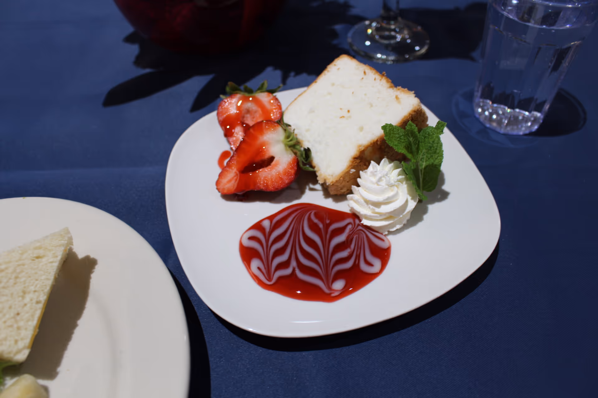 A dessert plate with a slice of white cake, two halved strawberries, a swirl of whipped cream garnished with a mint leaf, and a decorative red and white sauce on a white plate placed on a dark blue tablecloth. A glass of water and part of another plate with a sandwich are also visible.
