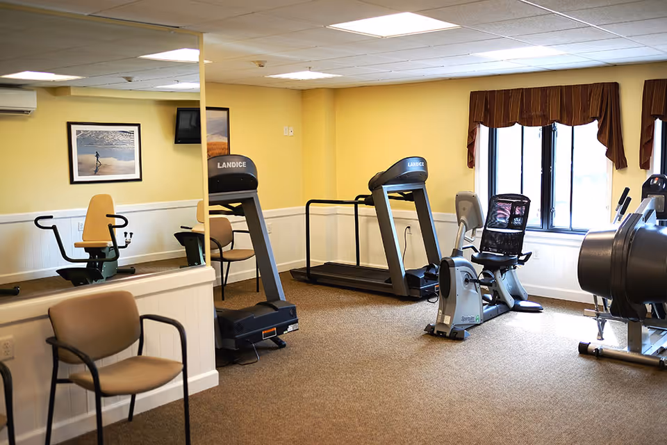 A small fitness room with exercise equipment including a treadmill, stationary bike, and weight machine. The room has beige walls, a large mirror on one wall, a window with brown valances, and a framed picture on the wall. There are also a couple of chairs placed near the mirror.