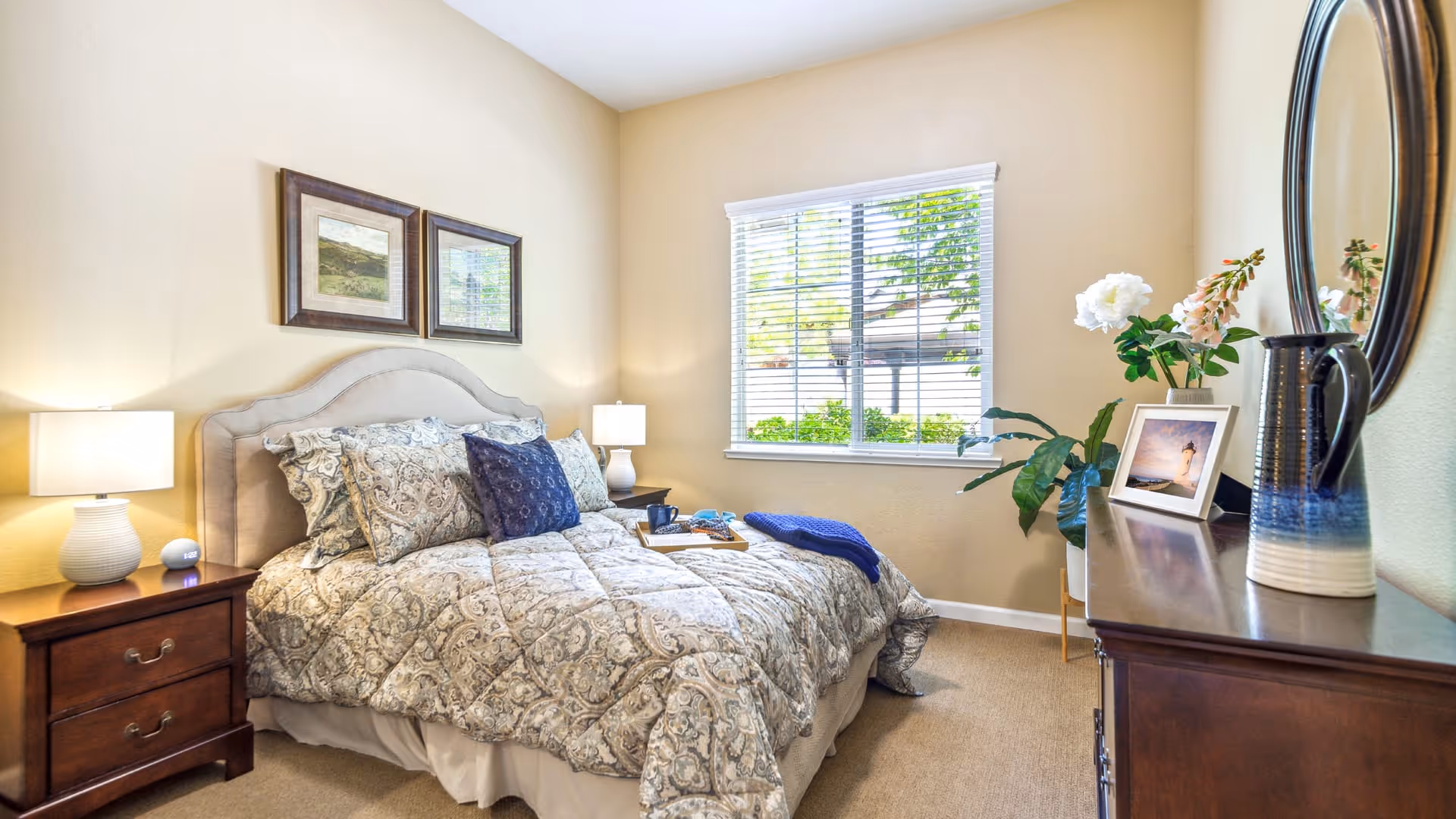 A cozy bedroom with a large bed covered in patterned bedding and multiple pillows. There are two framed pictures on the wall above the bed. On either side of the bed are wooden nightstands with lamps. A window with white blinds lets in natural light, and a wooden dresser with a round mirror, a vase with flowers, and a framed photo is on the right side of the room.