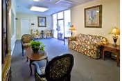 A well-lit living room area with floral patterned sofa and chairs, a wooden coffee table with a green plant, side tables with lamps, framed artwork on the walls, and large windows letting in natural light.