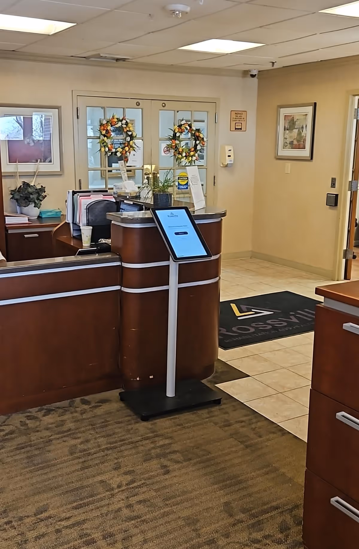 Reception area of Rossville Rehabilitation and Healthcare Center with a wooden front desk, a digital check-in kiosk, decorative wreaths on glass double doors, framed artwork on the walls, and a floor mat with the facility's name.