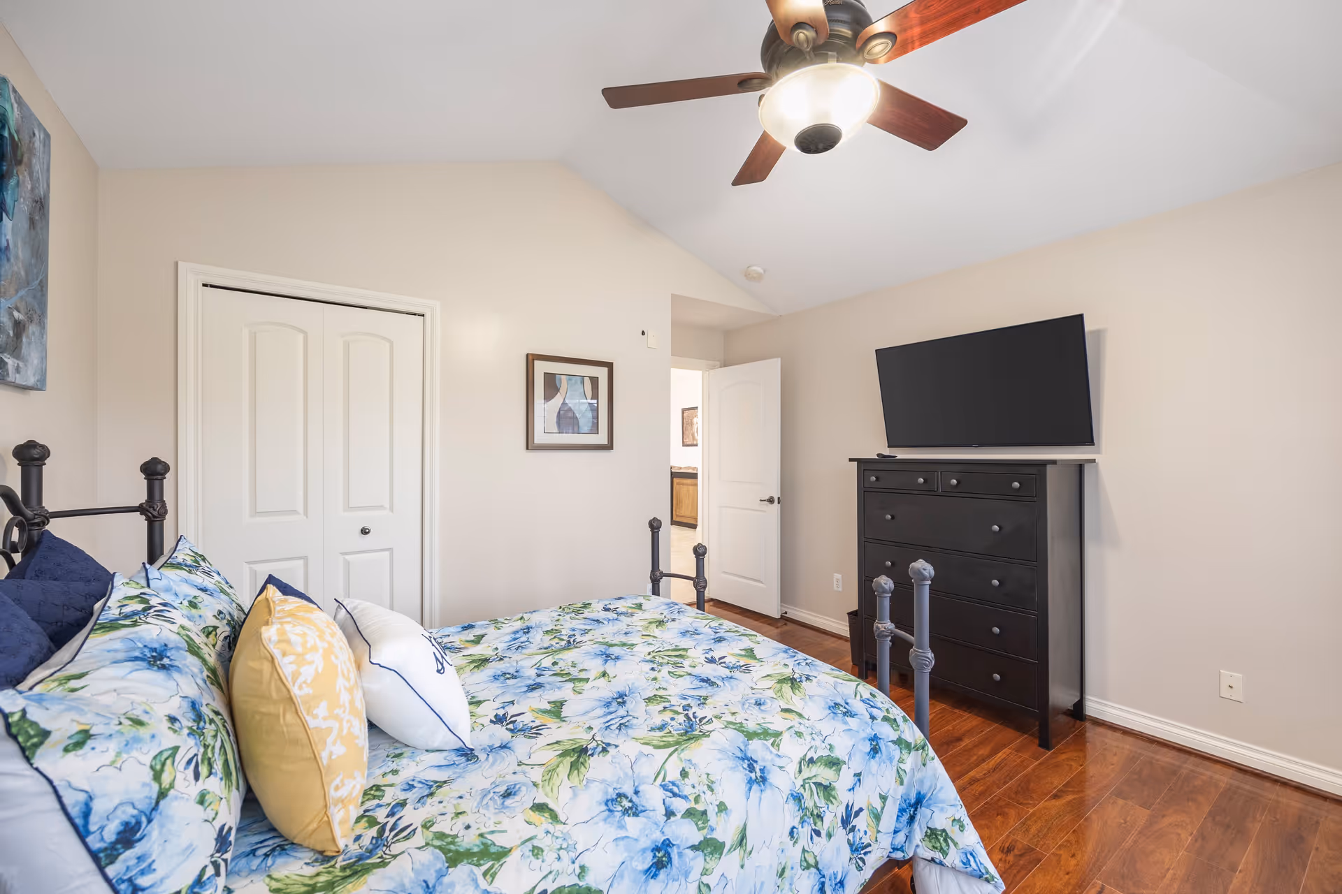 Bedroom with floral bedding, a ceiling fan, a black dresser with a TV, and a partially open door.