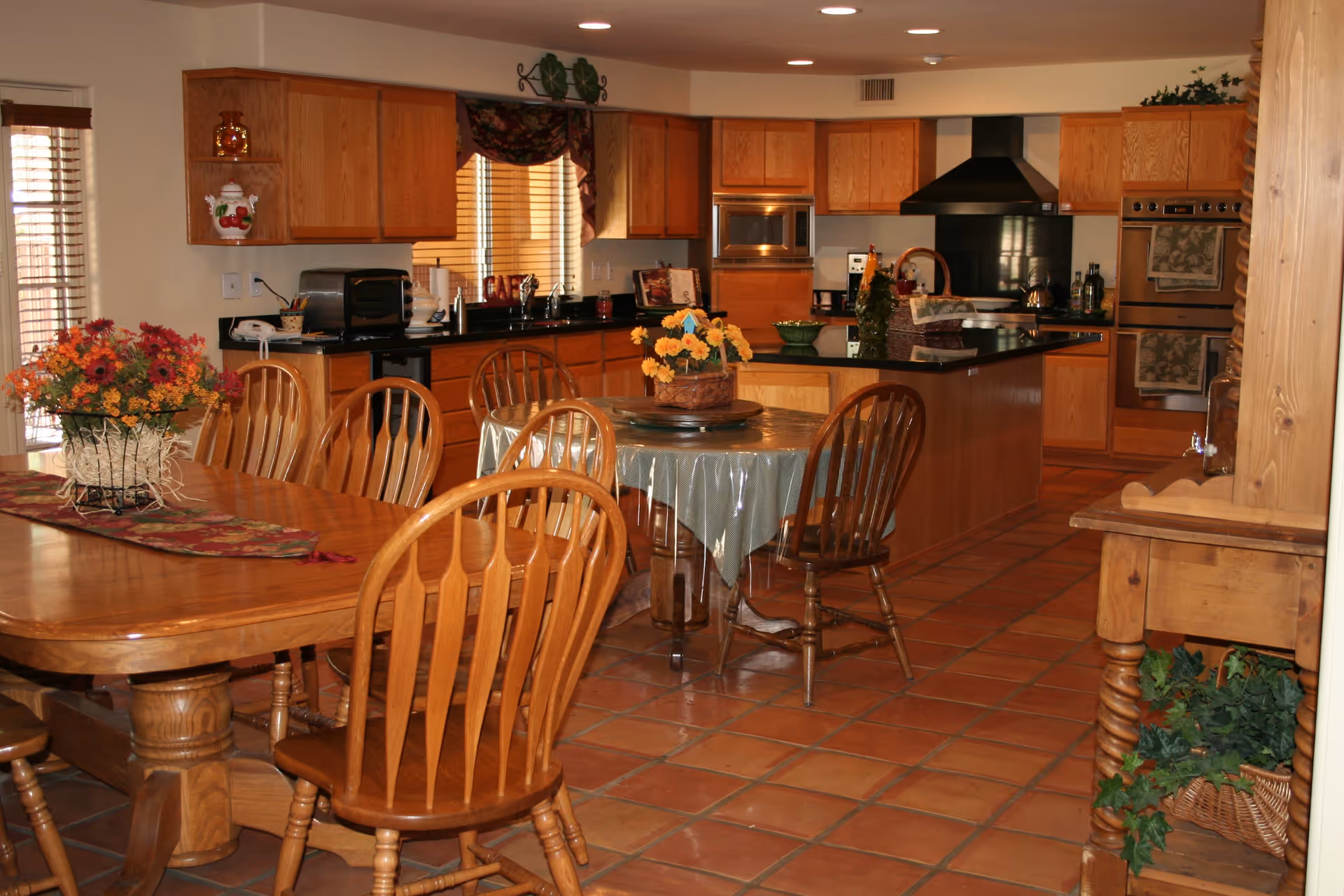 A warm, inviting kitchen and dining area with wooden cabinets, a black countertop, and terracotta tile flooring. The kitchen features a stove with a black range hood, built-in oven, microwave, and various kitchen appliances. There is a wooden dining table with chairs and a smaller round table with a floral centerpiece. The room is decorated with flowers and plants, and natural light filters through the window blinds.