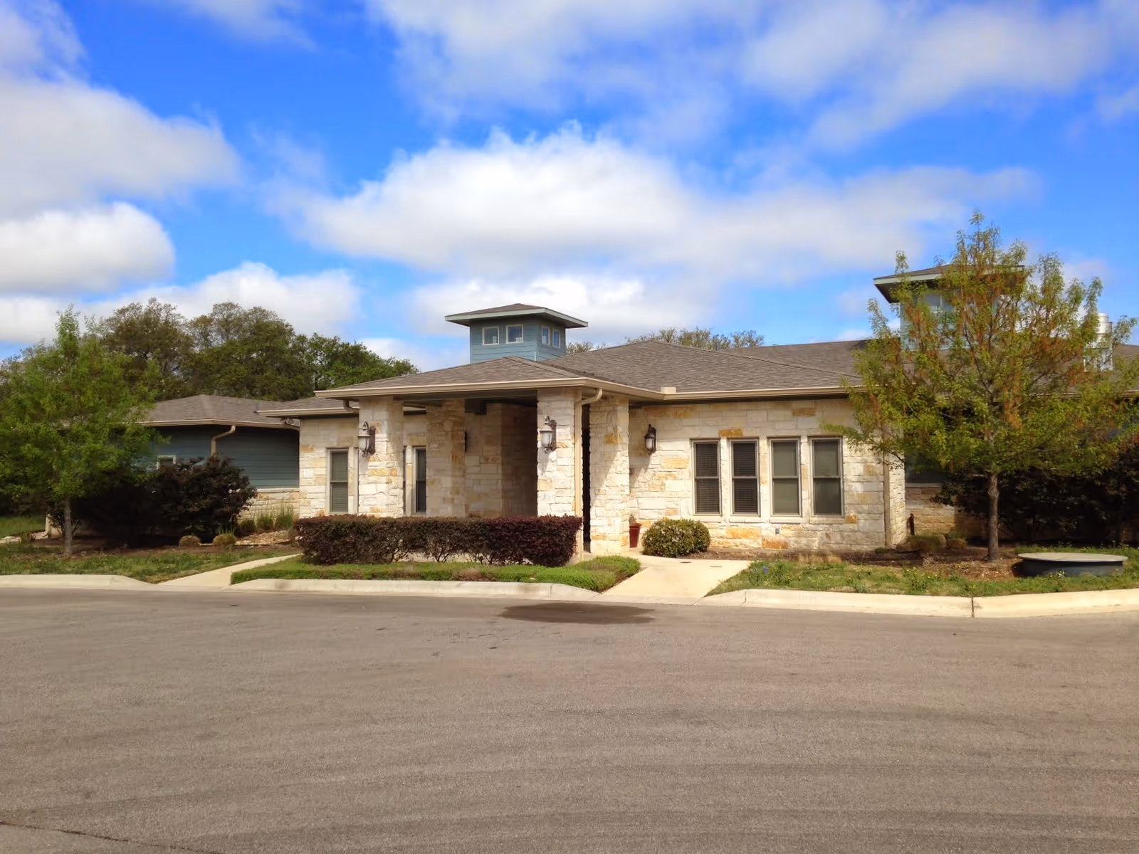 Exterior view of a single-story stone building with a covered entrance, surrounded by trees and shrubs under a partly cloudy blue sky.
