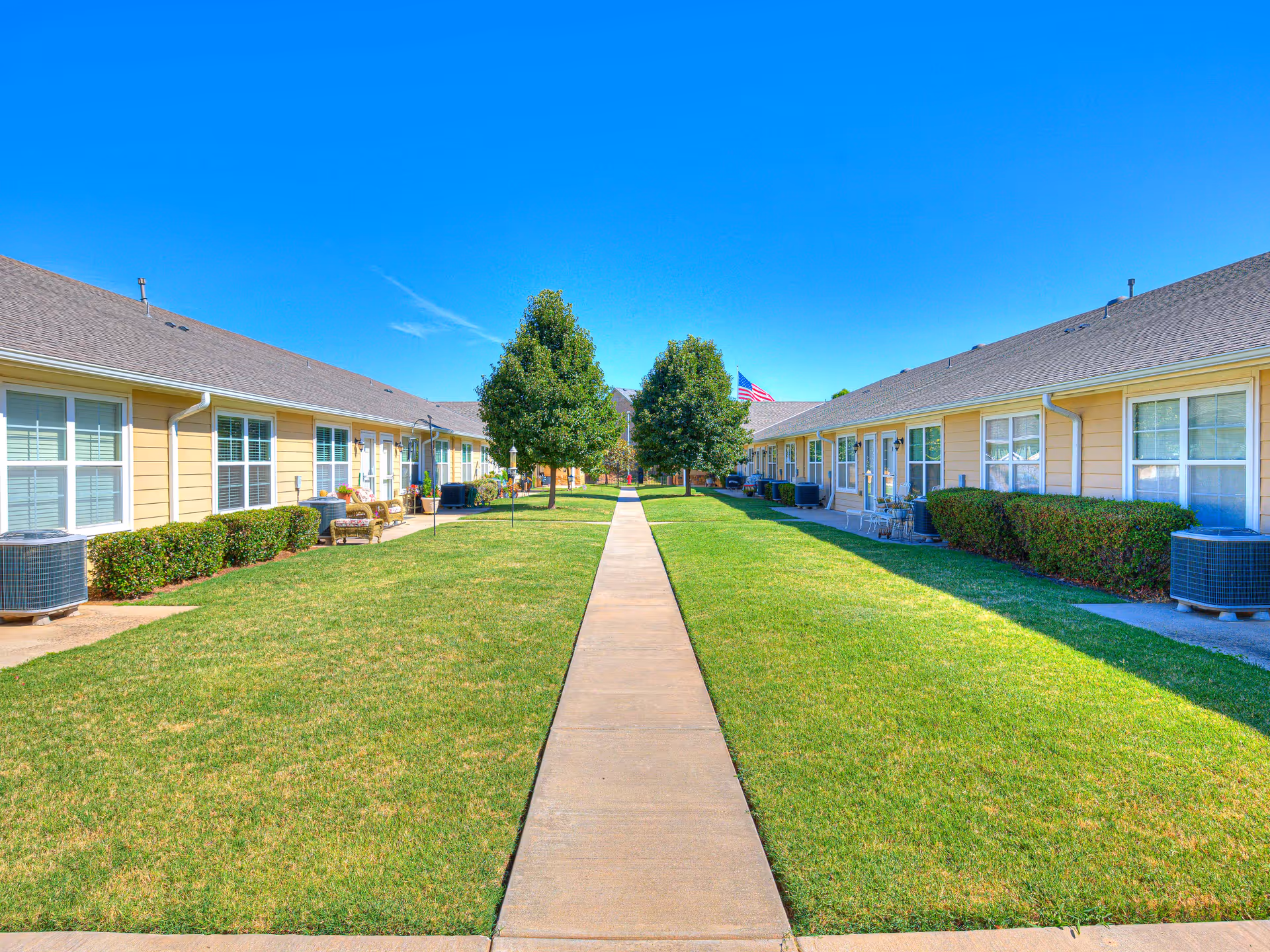Sunlit outdoor courtyard between beige senior living buildings with a central path and trees