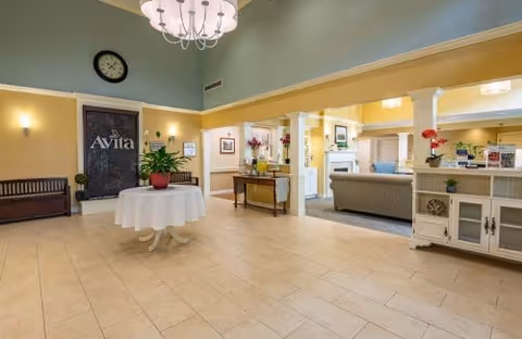 Bright lobby of Avita of Brunswick with a chandelier, round table with a potted plant, bench seating, and an open seating area under a high ceiling.