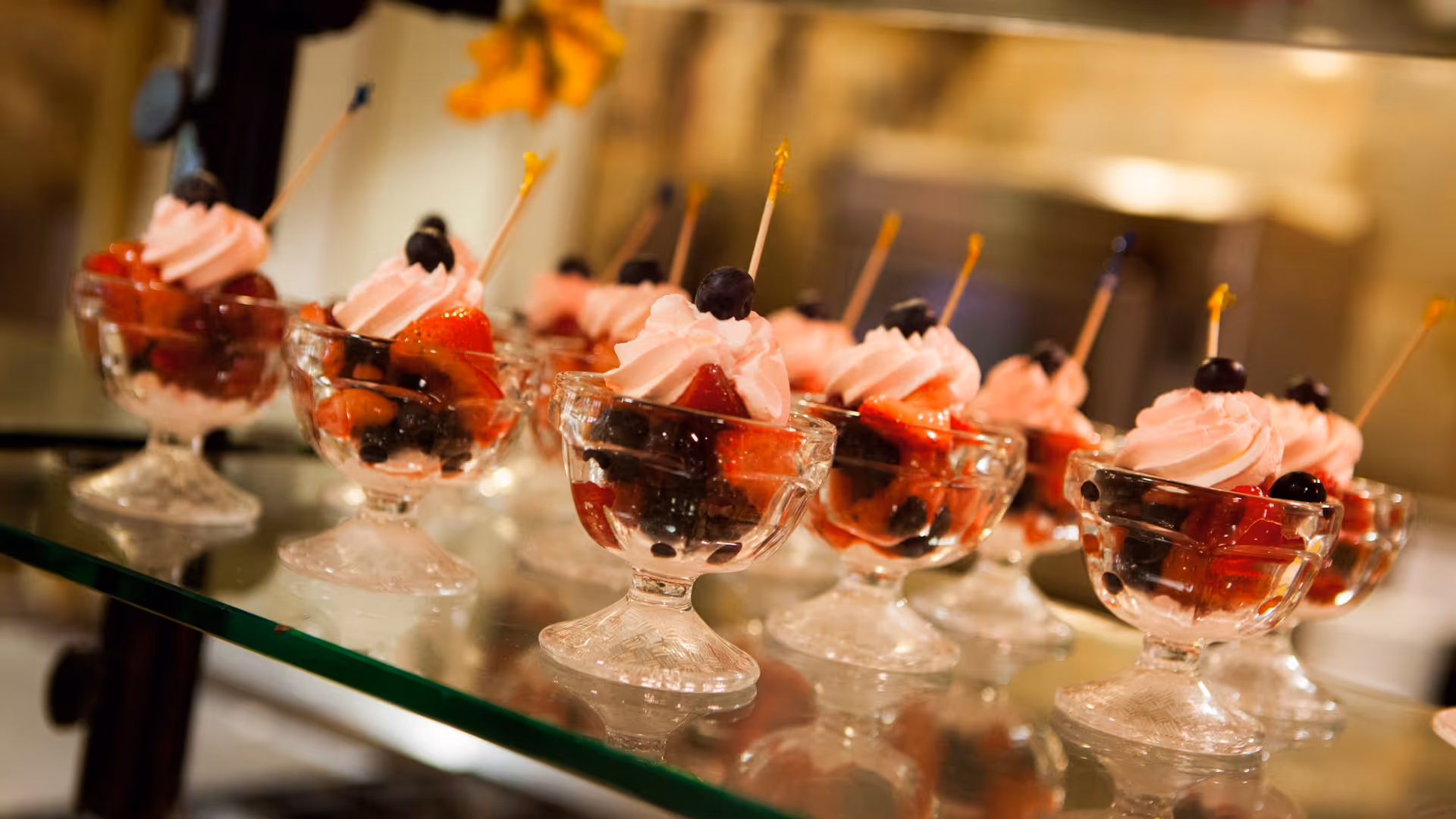 Close-up view of multiple small glass dessert cups filled with mixed berries and topped with pink whipped cream and a single blueberry on a toothpick, arranged on a glass shelf.