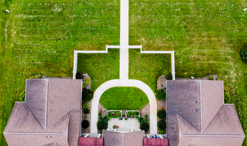 Aerial view of a senior living facility showing two buildings with brown roofs separated by a curved concrete walkway and green lawn. The walkway leads to a straight path extending upwards, bordered by white fencing and landscaped with bushes and flower beds.