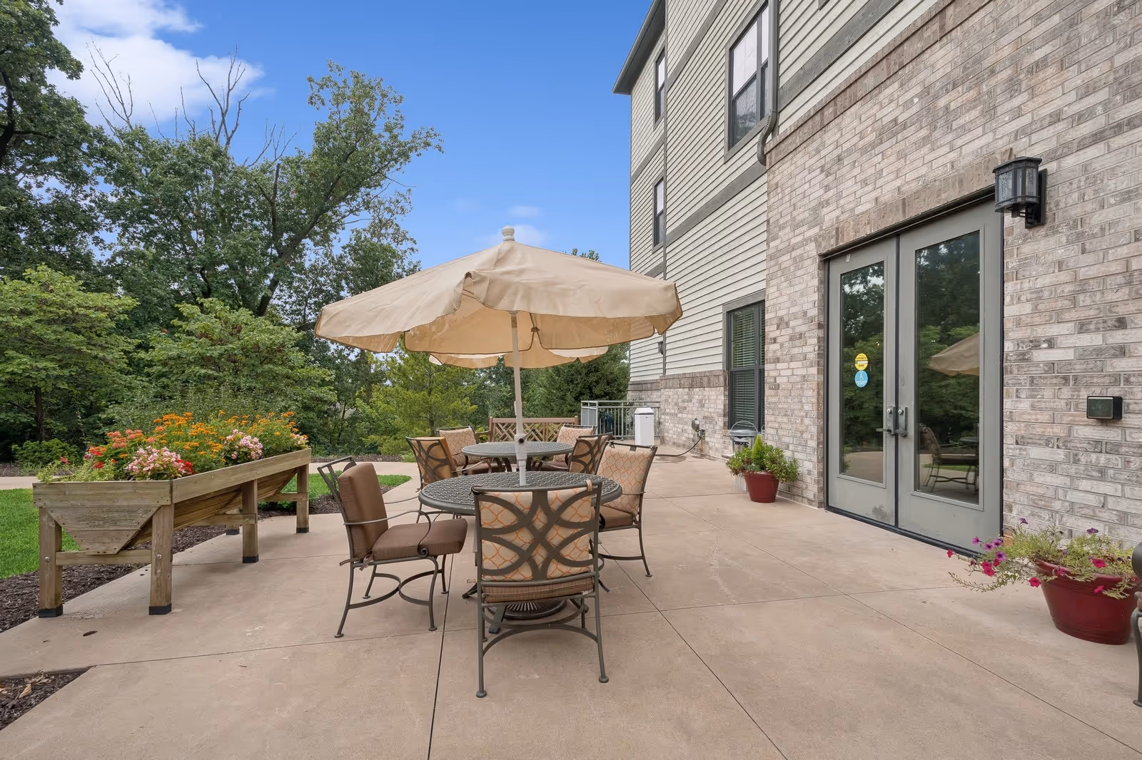 Outdoor patio area at The Grande at Laumeier Park featuring a round metal table with a beige umbrella and six cushioned chairs. There are flower planters with colorful flowers on the concrete patio, and the building exterior is visible with brick and siding walls and glass double doors. Trees and greenery surround the patio under a blue sky with some clouds.