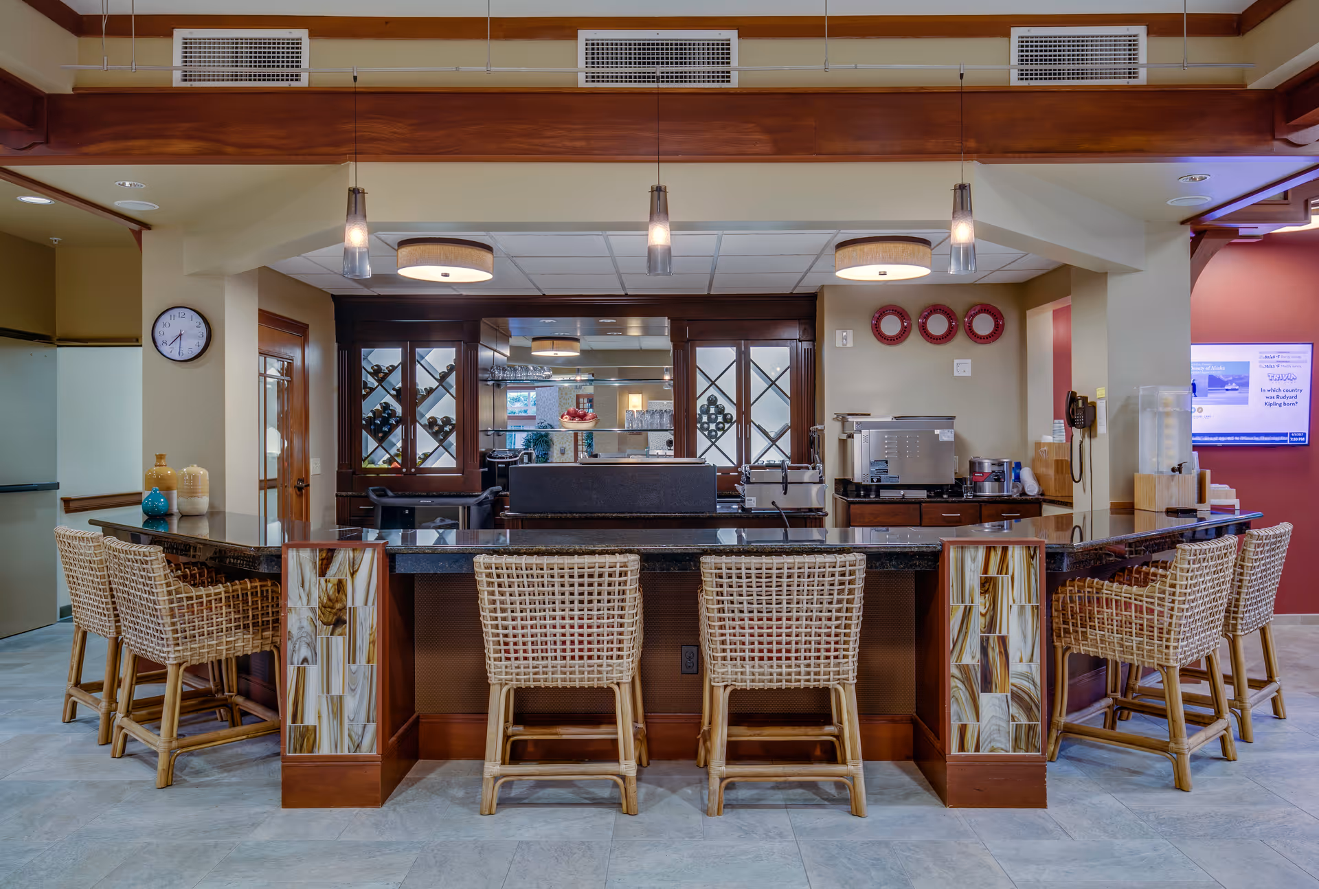 Communal bar area with a granite countertop, wicker barstools, pendant lights, and cabinetry in a senior living facility.