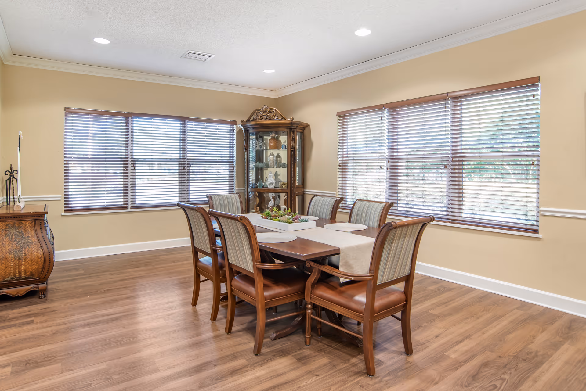 A dining room with a wooden table set for six, featuring cushioned chairs with striped backs and leather seats. The room has large windows with wooden blinds, light beige walls, and wood flooring. There is a decorative wooden cabinet with glass doors in the corner displaying various decorative items.