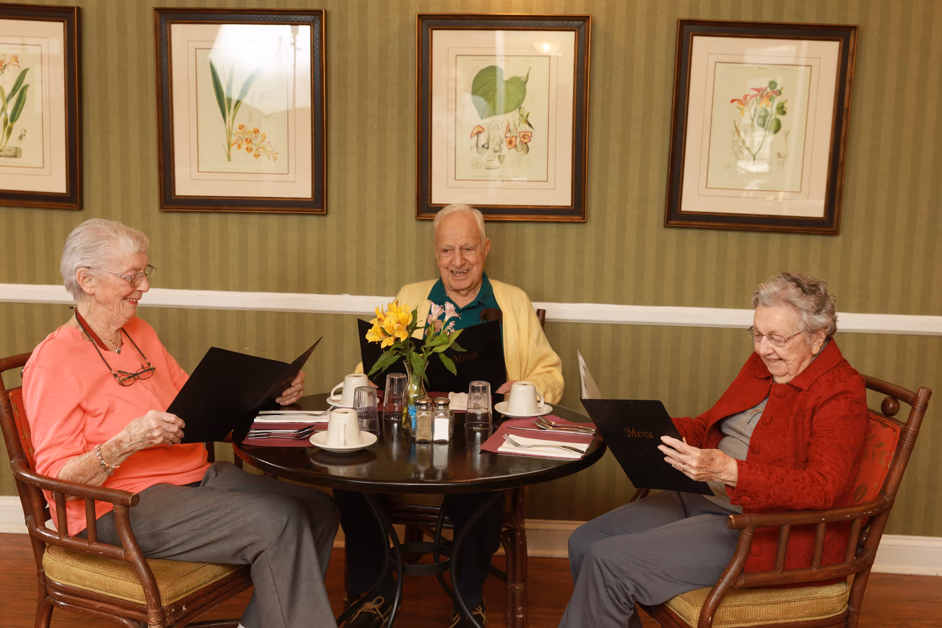 Three elderly individuals sitting around a round dining table in a room with green striped wallpaper and framed botanical prints. They are looking at menus and appear to be in a dining setting with cups, glasses, and a vase with yellow flowers on the table.