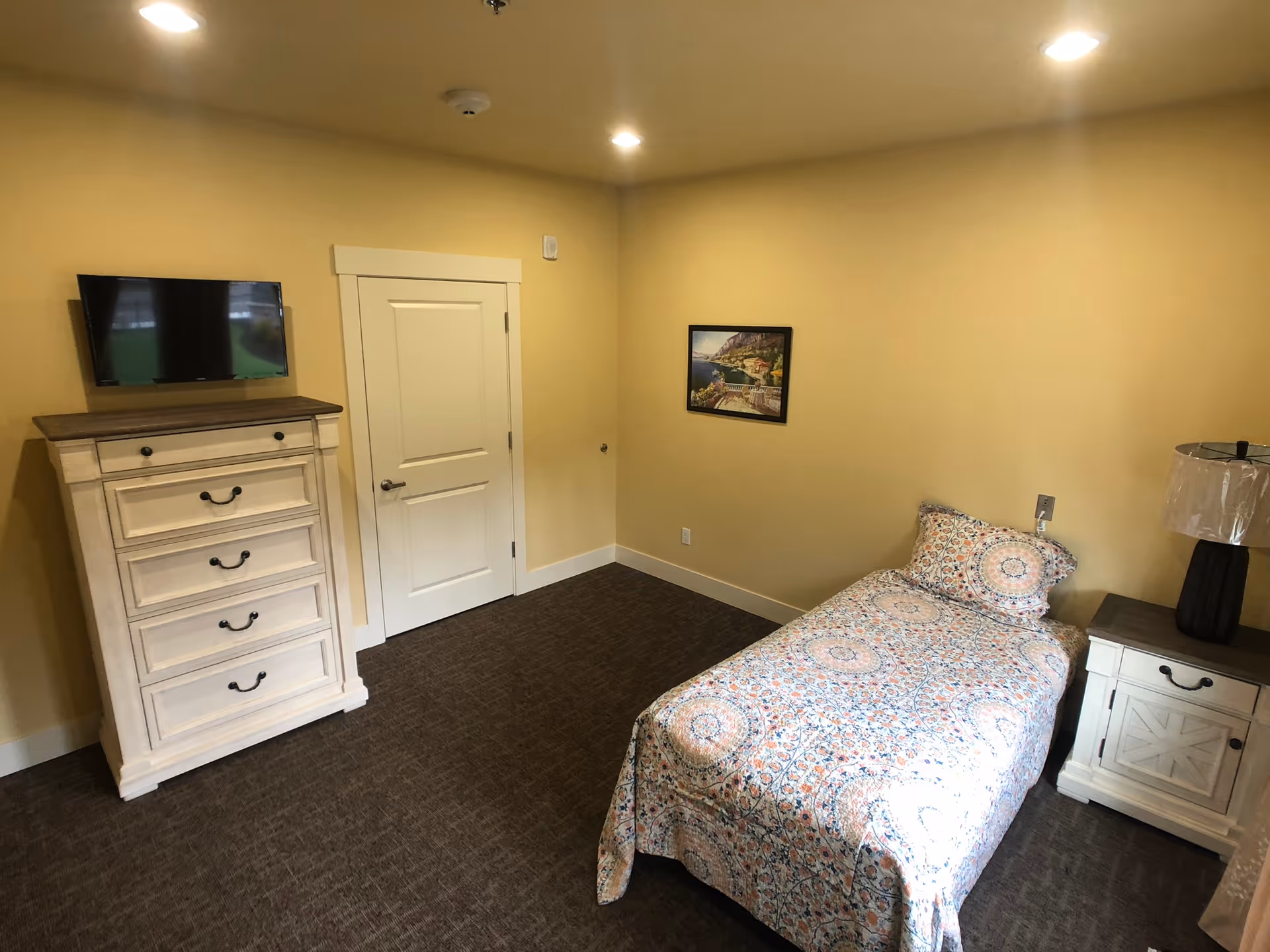 A small bedroom with beige walls and carpeted floor. The room contains a single bed with a colorful patterned bedspread and matching pillow, a white nightstand with a lamp, a white dresser with a dark wooden top, and a wall-mounted flat-screen TV. A framed picture hangs on the wall above the bed.