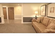 A waiting area in a senior living facility with a striped sofa, a side table with a lamp, and two framed pictures on the wall. The floor is carpeted and there are doors and a hallway visible in the background.