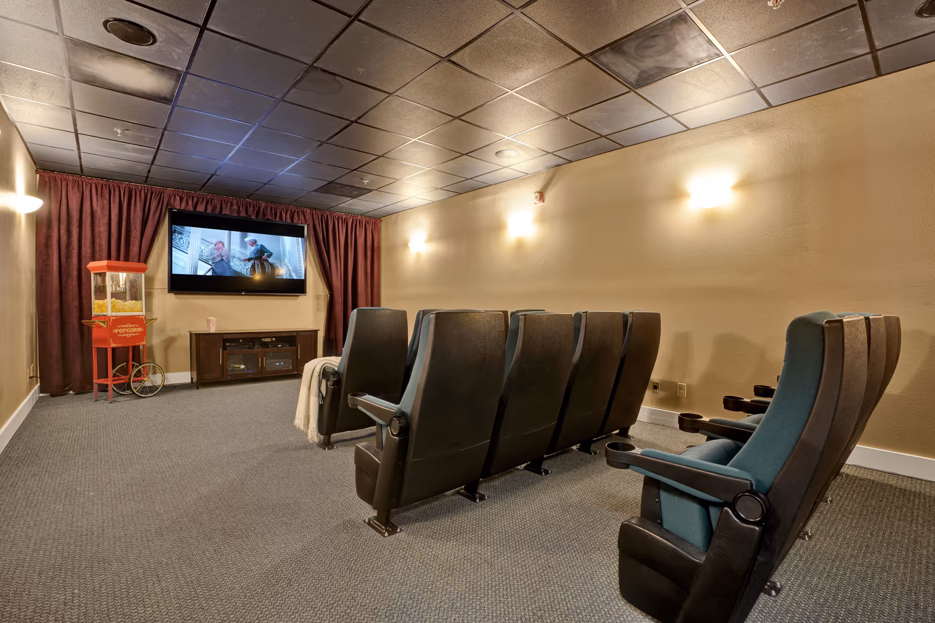 A small movie theater room with two rows of black theater seats facing a wall-mounted flat screen TV. There is a red popcorn machine on the left side near the TV, and the walls are painted beige with soft wall lights. The ceiling has black tiles, and the floor is carpeted in gray.