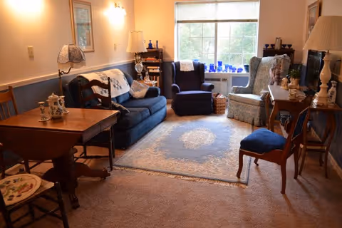 A cozy living room with a blue sofa, two armchairs, a wooden table with chairs, a patterned area rug, and various decorative items including lamps, framed pictures, and blue glassware on the windowsill.