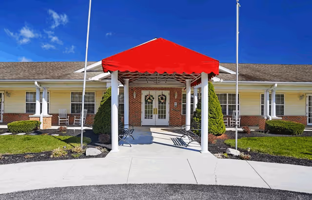 Front entrance of a single-story senior living facility with a red canopy over the walkway, white pillars, and double glass doors decorated with wreaths. The building has yellow siding with brick accents and is surrounded by neatly trimmed bushes and a clear blue sky.