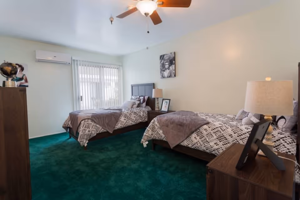 A bedroom with two twin beds, each with patterned bedding and a gray throw blanket. There are two nightstands with lamps and framed photos, a dresser with a globe on top, a ceiling fan with a light, and vertical blinds covering a window or sliding door. The room has green carpet and light-colored walls.
