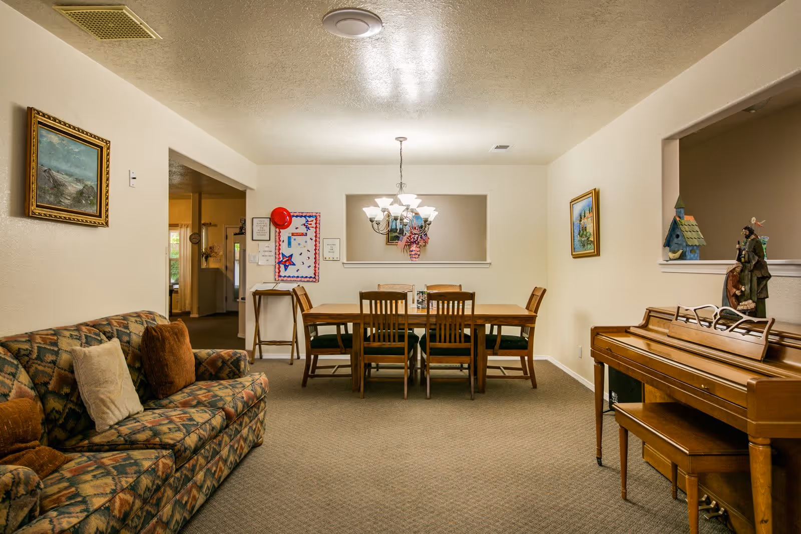 Interior view of a senior living facility room with a patterned sofa on the left, a wooden dining table with six chairs in the center under a chandelier, and a wooden piano with a bench on the right. The walls are decorated with framed paintings and small decorative items. The room has beige carpet and light-colored walls.