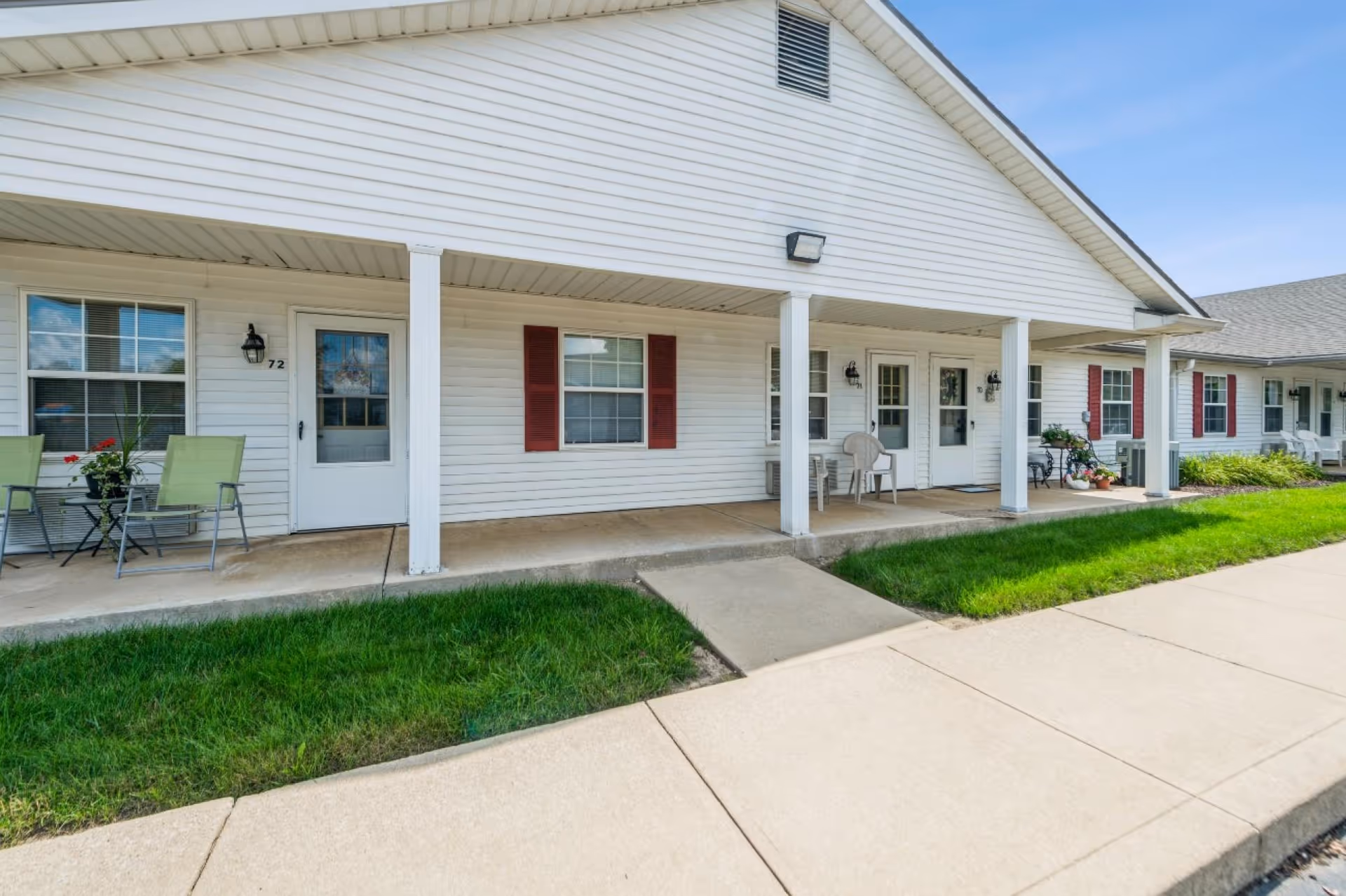 Exterior view of a single-story residential building with white siding and red shutters. The building has a covered porch supported by white columns, with several doors and windows visible. There are chairs and small tables with plants on the porch, and a concrete sidewalk and green grass in front.