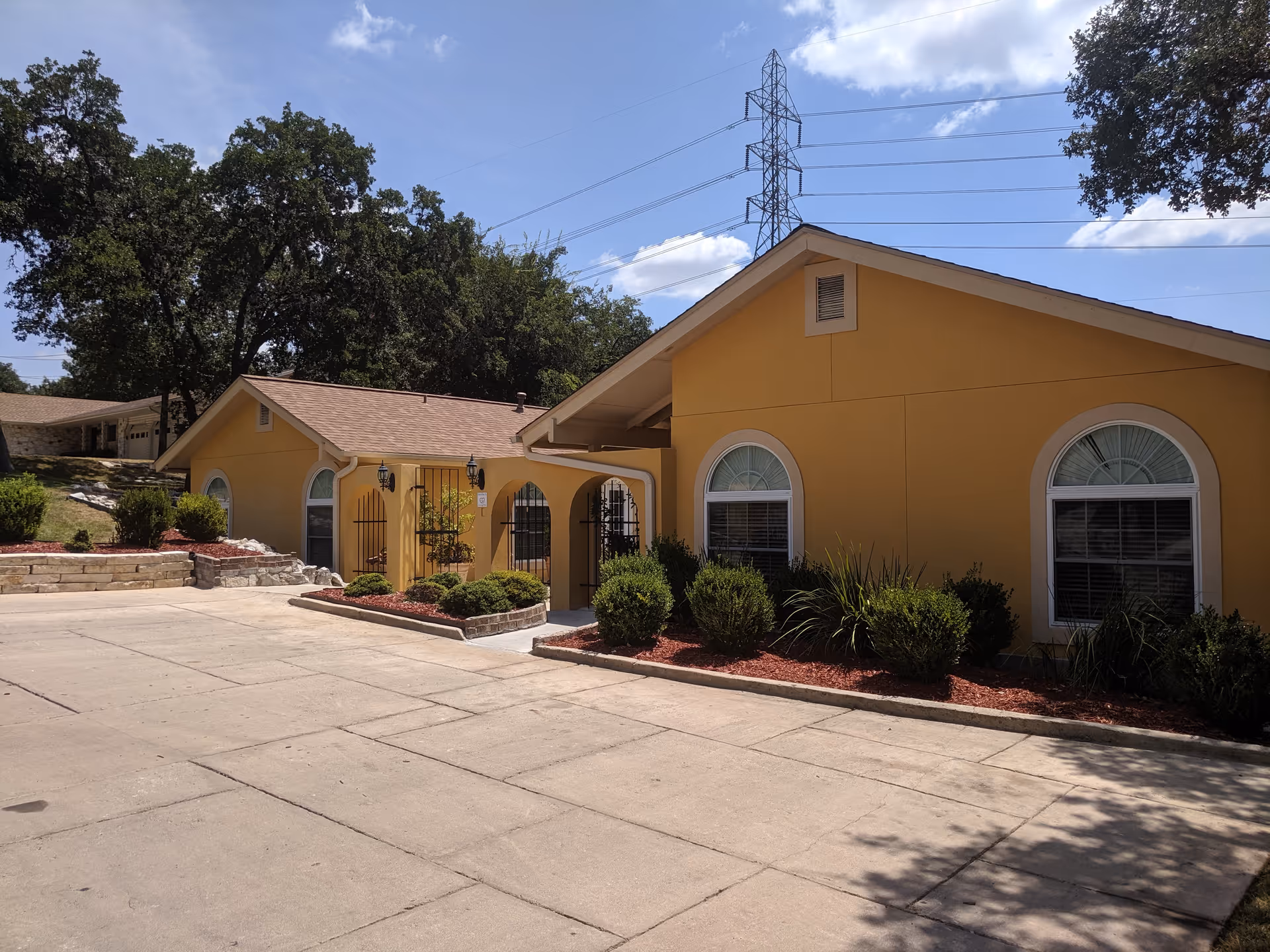 Single-story yellow building with arched windows, landscaped shrubs and a paved driveway under a blue sky with power lines overhead.