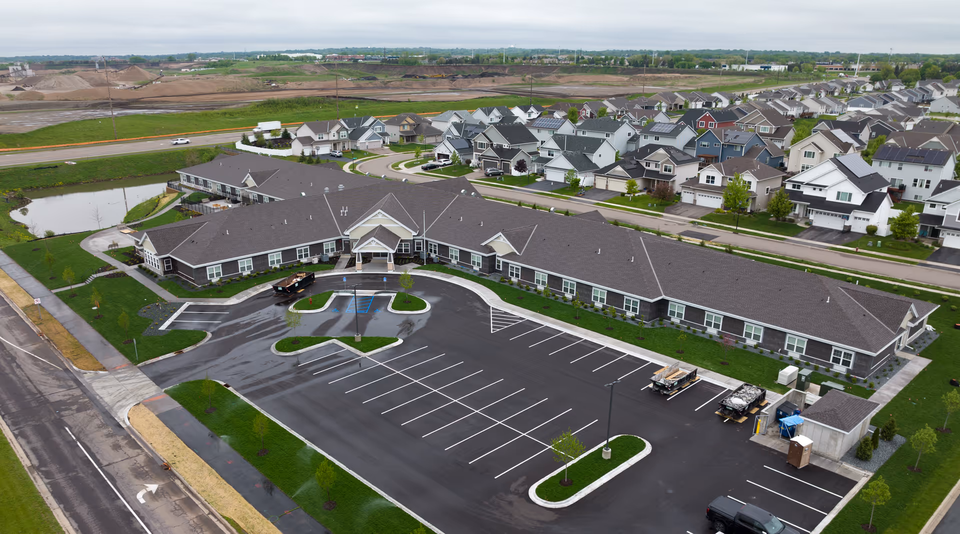 Aerial view of Boden Senior Living facility showing a single-story building with a large parking lot in front, surrounded by green lawns and a residential neighborhood with many houses in the background under a cloudy sky.