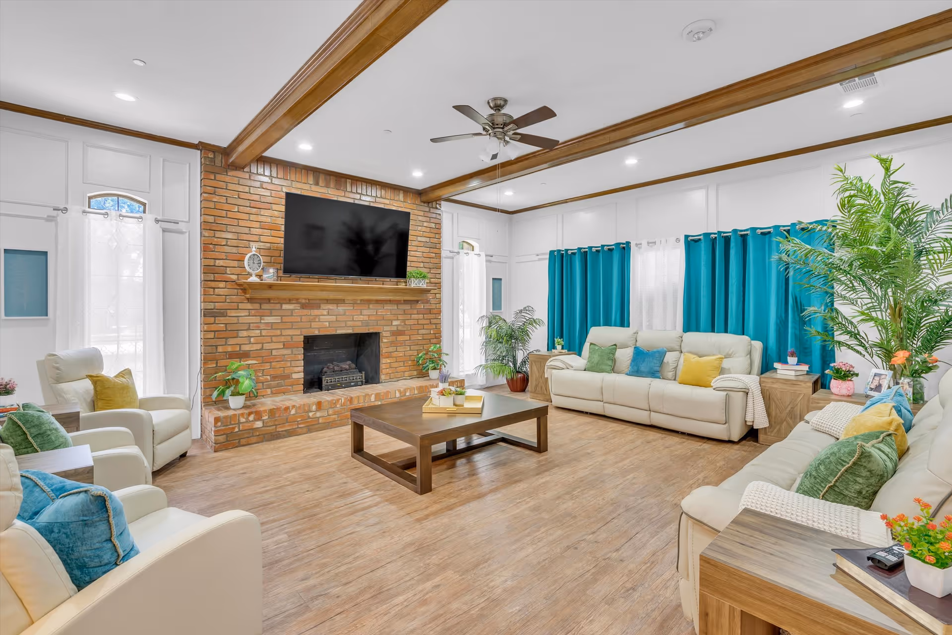 Bright, spacious living room with sofas and armchairs, a brick fireplace with mounted TV, coffee table, and teal curtains.