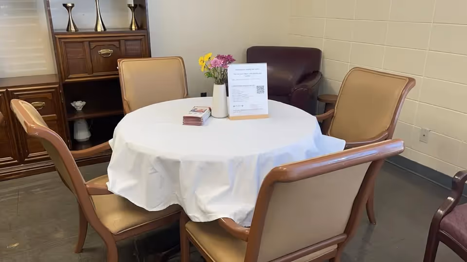 Round table with a white tablecloth, vase of flowers and pamphlets surrounded by four chairs in a small common room.
