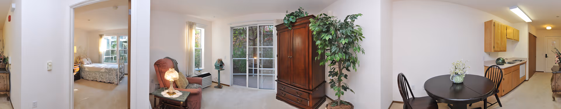 Panoramic interior view of an apartment showing a living area with seating and an armoire, sliding glass doors, an adjoining bedroom, and a small dining area with kitchenette.