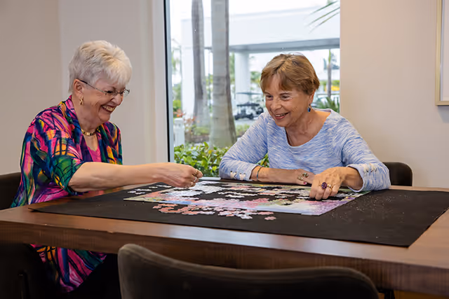 Two elderly women sitting at a table indoors, smiling and working together on assembling a jigsaw puzzle. There is a large window behind them showing some outdoor greenery and a golf cart.