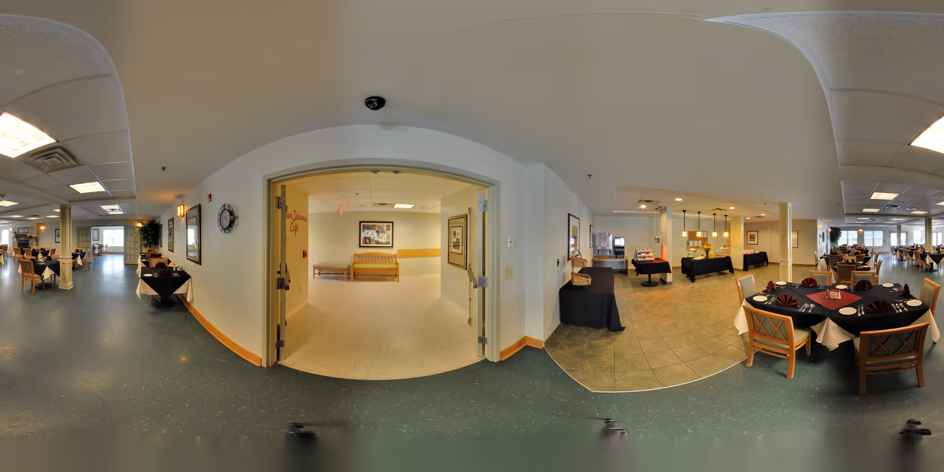 Panoramic view of a senior living facility dining area with multiple tables set with black and burgundy tablecloths, napkins, and place settings. To the right, there is a buffet area with food warmers and beverage dispensers. In the center, double doors lead to a hallway with a bench and framed pictures on the walls. The space is well-lit with ceiling lights and has a mix of tiled and vinyl flooring.