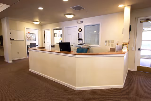 Reception desk area inside a senior living facility with a computer, office chair, and paperwork. The walls are light-colored, and there are doors and framed pictures in the background. The floor is carpeted, and ceiling lights illuminate the space.