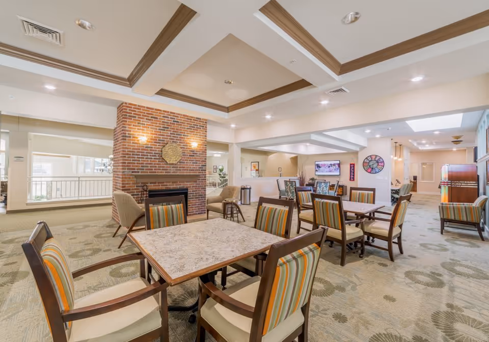 A spacious and well-lit common area with multiple tables and chairs featuring striped upholstery. The room has a carpeted floor with a patterned design, a brick fireplace with two wall sconces, and a large clock on the far wall. There is a television mounted on the wall and a jukebox in the background. The ceiling has recessed lighting and decorative beams.