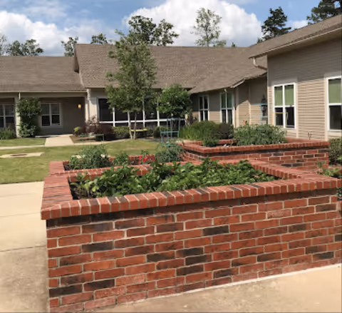 Outdoor view of a senior living facility with raised brick garden beds filled with plants and greenery. The building has beige siding and multiple windows, surrounded by a well-maintained lawn and trees under a partly cloudy sky.
