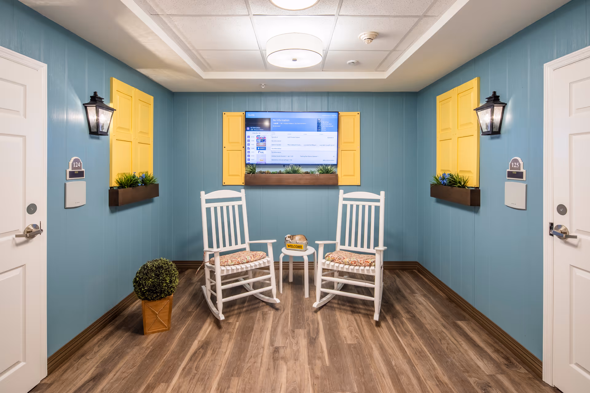 A cozy interior sitting area with two white rocking chairs with patterned cushions facing each other, separated by a small round table with a welcome sign and a decorative dog figurine. The walls are painted blue with yellow window shutters and flower boxes beneath the windows. Two white doors labeled 124 and 125 flank the sitting area, which has wood flooring and wall-mounted lantern-style lights.