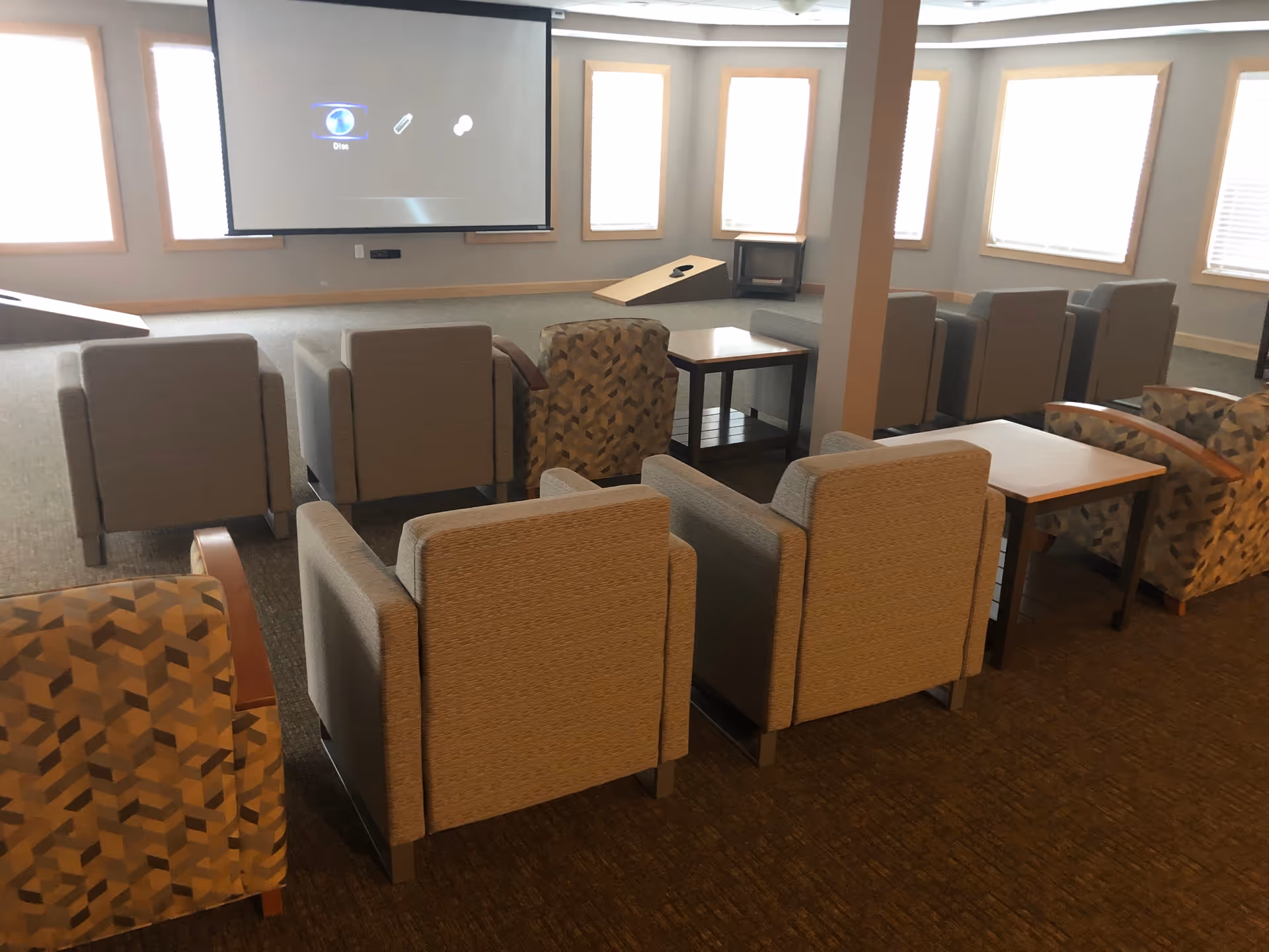 A senior community common room with multiple armchairs arranged in rows facing a large projector screen. The room has several windows with blinds and a carpeted floor. There are small tables placed between some of the chairs.