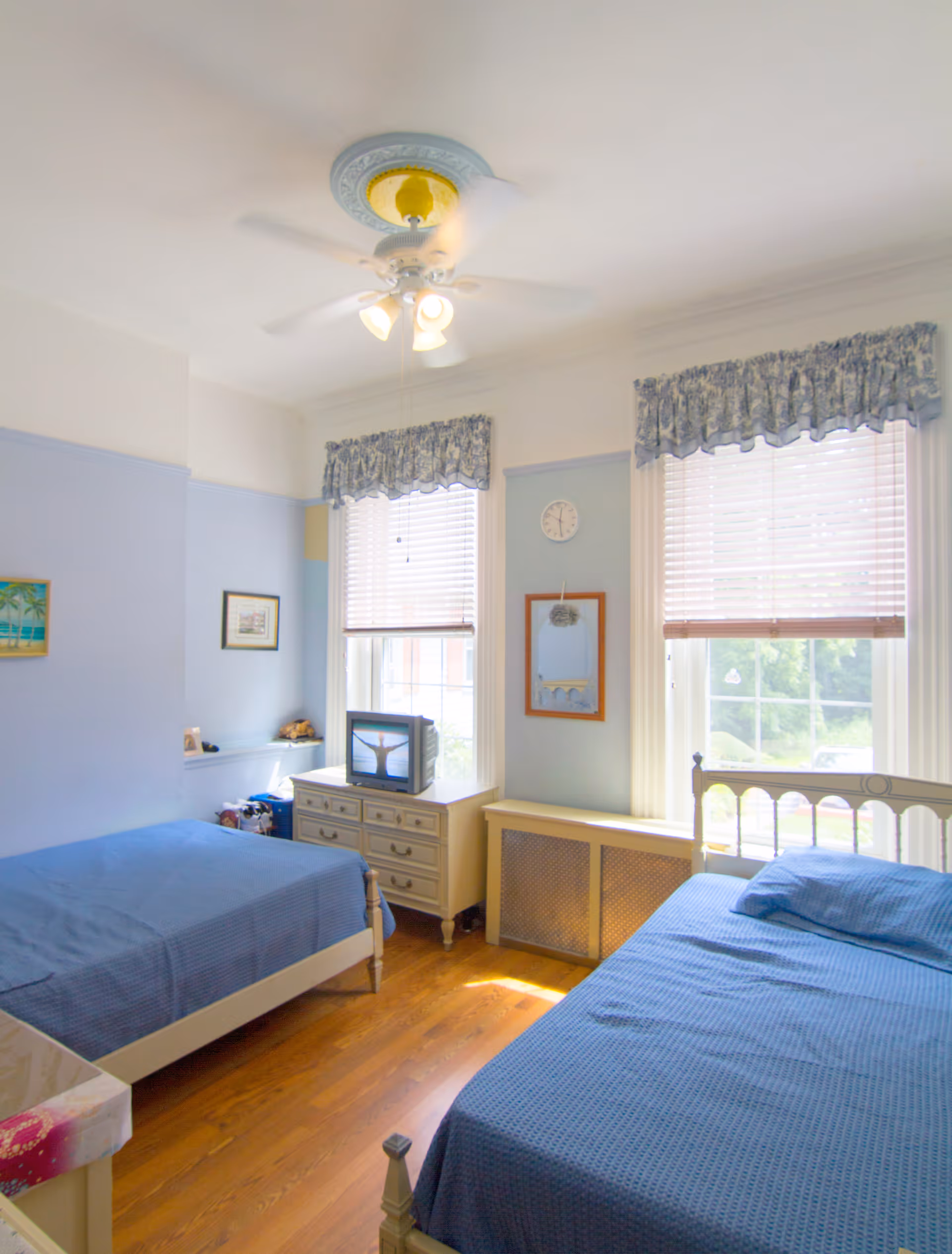 Sunlit bedroom with two twin beds in blue bedding, a dresser with a small TV, and two large windows with blinds.
