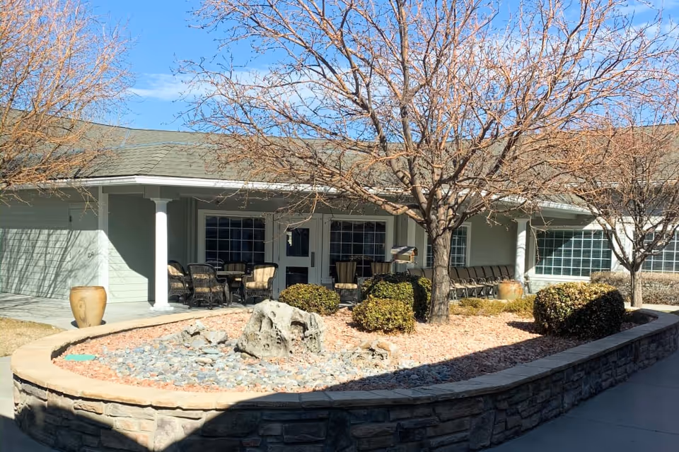 Outdoor courtyard area of Aspen Ridge Alzheimer’s Special Care Center featuring leafless trees, trimmed bushes, a stone-bordered garden bed with rocks, and a covered patio with chairs and tables.
