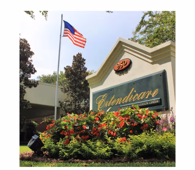 Outdoor view of the Extendicare Health Center sign surrounded by green bushes and red flowers, with an American flag on a flagpole in the background and trees around.