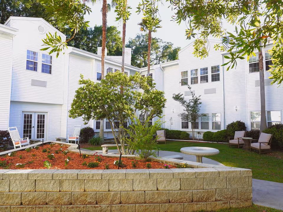 Outdoor courtyard area of a senior living facility with white two-story buildings surrounding a garden bed with small plants and trees. There are several chairs and small tables arranged on the grass and paved walkways, with tall palm trees and other greenery providing shade.