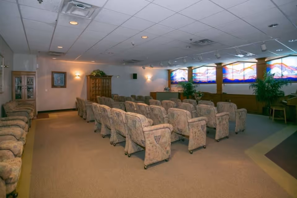 A room with multiple rows of cushioned armchairs arranged facing a podium or stage area. The room has a carpeted floor, beige walls, and a ceiling with recessed lighting. There are large stained glass windows on one side and some potted plants near the front. Wooden cabinets and framed artwork are visible along the walls.