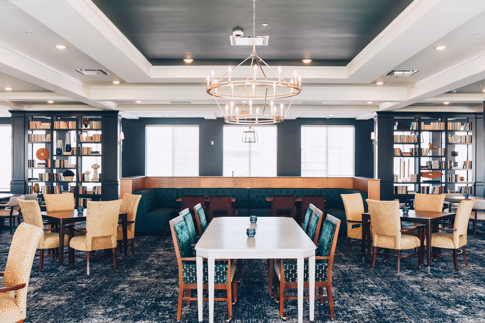 A spacious and elegant dining room with multiple tables and chairs arranged neatly. The room features a large chandelier hanging from a dark ceiling, large windows letting in natural light, and bookshelves filled with books and decorative items along the walls.