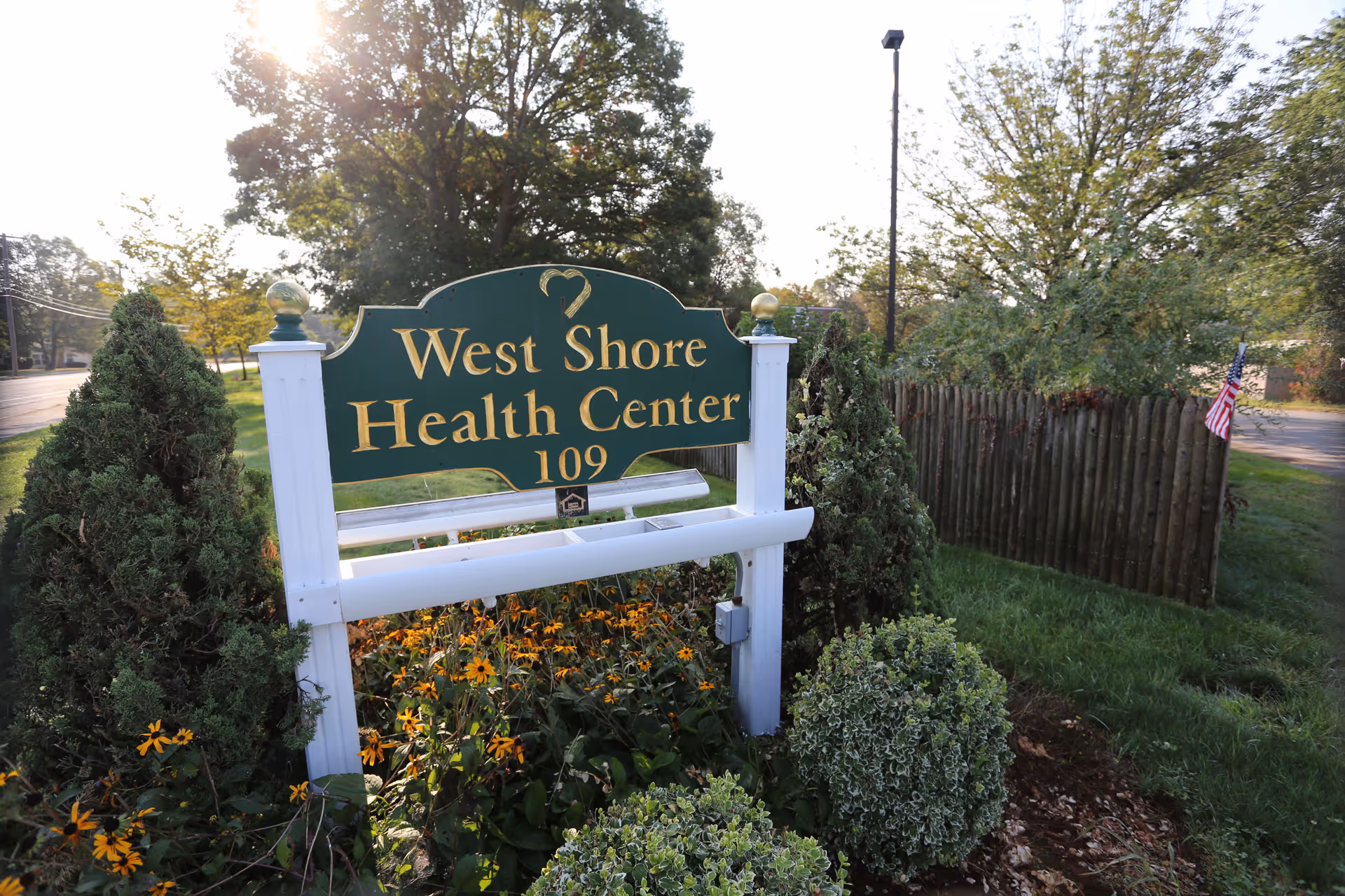 Green and gold sign reading "West Shore Health Center 109" surrounded by shrubs, flowers, and a small American flag at the facility entrance.