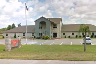 Exterior view of a single-story senior living facility building with a two-story central section, a flagpole with an American flag in front, a parking lot with a few cars, and a sign that reads 'Gulf Pointe Village'. The sky is partly cloudy.