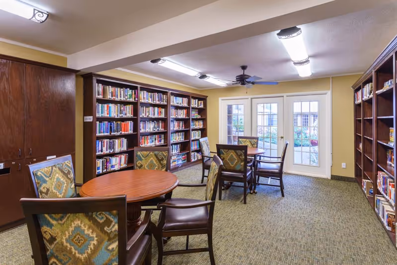 Small library/reading room with bookshelves, round tables, and upholstered chairs near French doors.